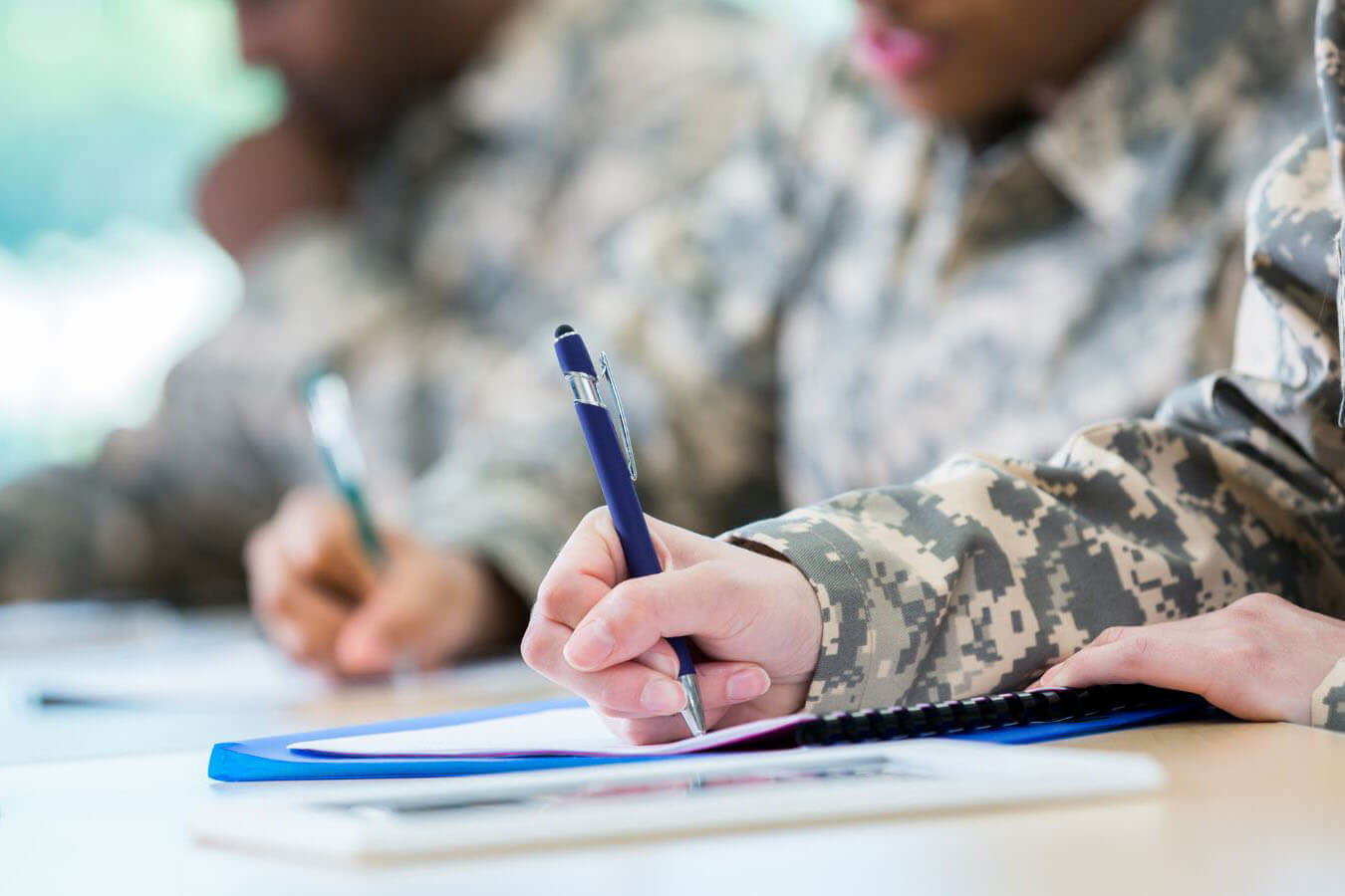 Veteran in military uniform seated at a desk, writing on a document with study materials nearby. Select 84 more words to run Humanizer.